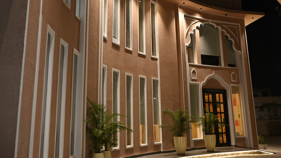 An evening view of the curved white exterior of Kadamb Kuteer, featuring multiple windows with warm interior lighting and a paved walkway.