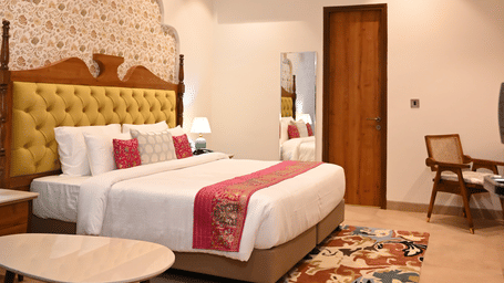 A guest room at Kadamb Kuteer with king bed, white linens, patterned pillows, a small seating area, and neutral-colored tiled flooring.