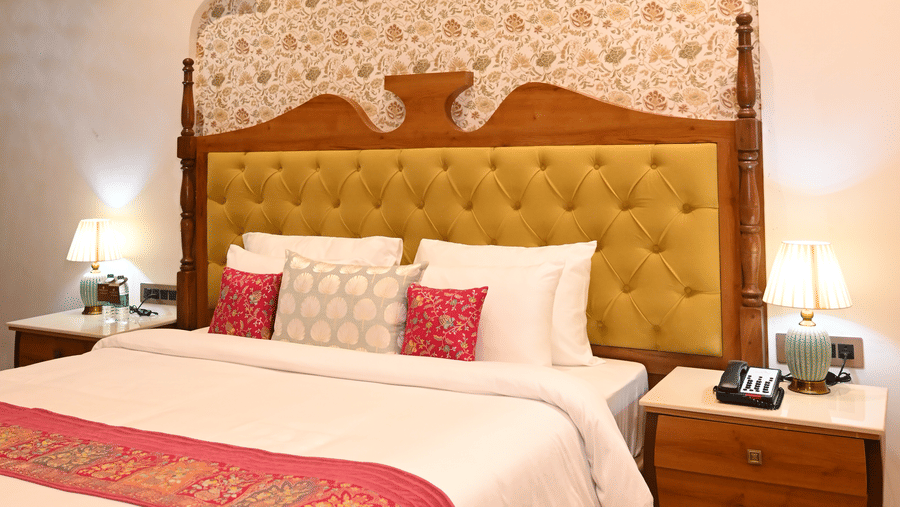 A bedroom at KAdamb Kuteer featuring a king sized bed with a yellow upholstered headboard, a wooden side table, and a window with light curtains.