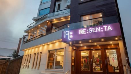 A low-angle shot of the Regenta Inn DLS, Rishikesh, at dusk. The building's illuminated entrance features a prominent purple RE:GEN:TA sign. Warm lights glow from inside, highlighting the glass doors and the steps leading up to the entrance. The sky behind