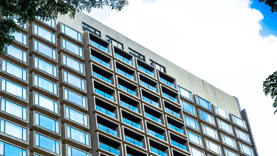 A tall modern hotel or office building with numerous reflective glass windows rises above leafy green tree branches.