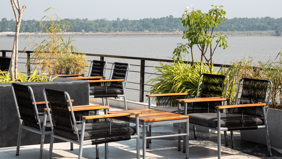 Outdoor seating area on a rooftop terrace, with tables and chairs near a railing, overlooking a large water body under a bright sky at Hotel Sonar Bangla, Taki.