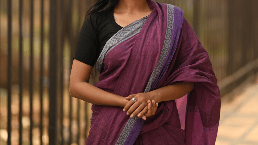 A woman posing for a picture in front of a metal gate and trees behind it, while wearing one of the popular Chettinad handloom sarees in purple colour.