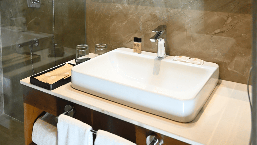A clean bathroom at Kadamb Kuteer with beige wall tiles, a white rectangular vessel sink on a wooden counter, and a large wall-mounted mirror.