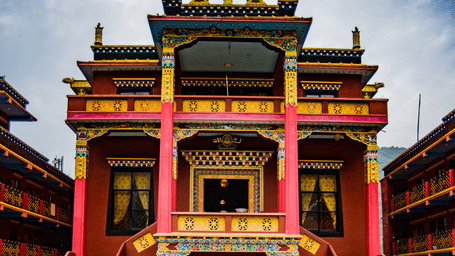 Ornate red and gold Tibetan Buddhist monastery courtyard with a large copper urn holding floating flowers, monks walking past, and a dog resting nearby.