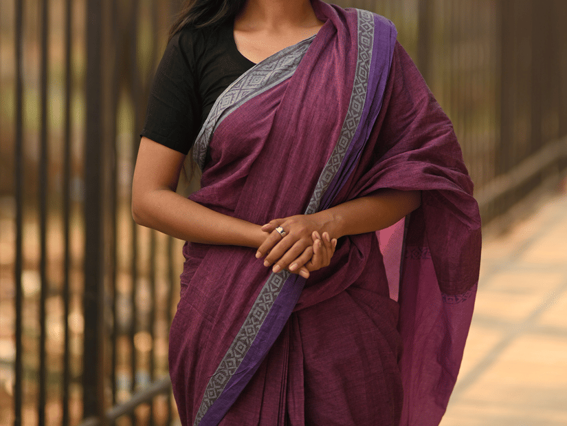 A woman posing for a picture in front of a metal gate and trees behind it, while wearing one of the popular Chettinad handloom sarees in purple colour.
