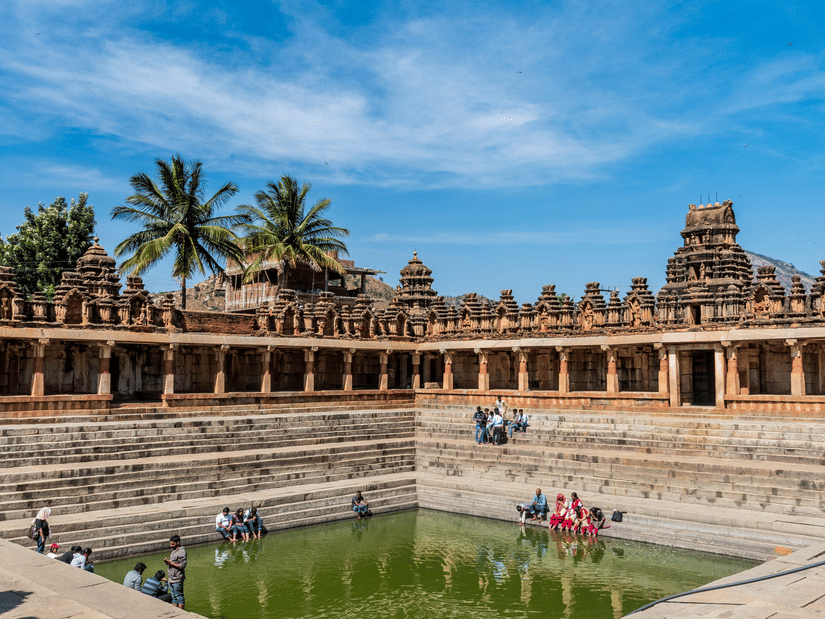 A large, rectangular stone stepped tank filled with water is surrounded by a pillared walkway and palm trees under a bright blue sky.