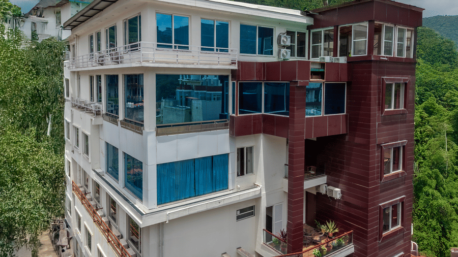 A vertical shot of the exterior of a multi-story hotel building with a mix of white and reddish-brown paneling. The building has large windows with blue tints and balconies with potted plants. It is set against a backdrop of green, forested hills under a c