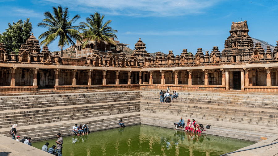 A large, rectangular stone stepped tank filled with water is surrounded by a pillared walkway and palm trees under a bright blue sky.
