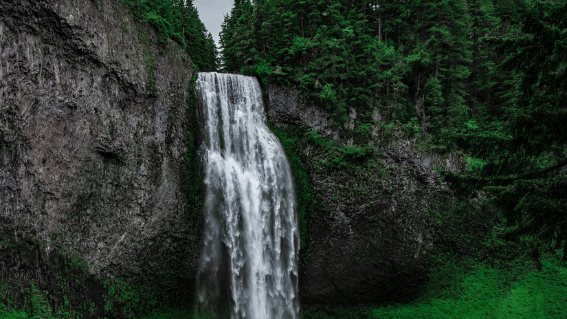 A view from below of Kune Waterfalls cascading from in between two peaks and numerous trees seen above them
