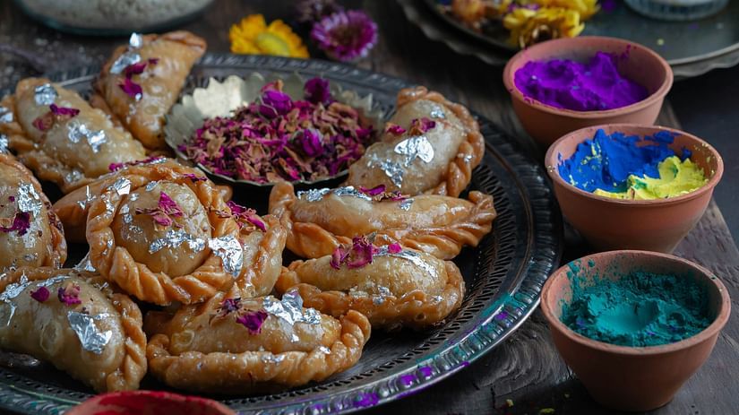 A plate of golden-brown Gujiya, a sweet Indian pastry, surrounded by bowls of colorful Holi powder and glasses of Thandai.