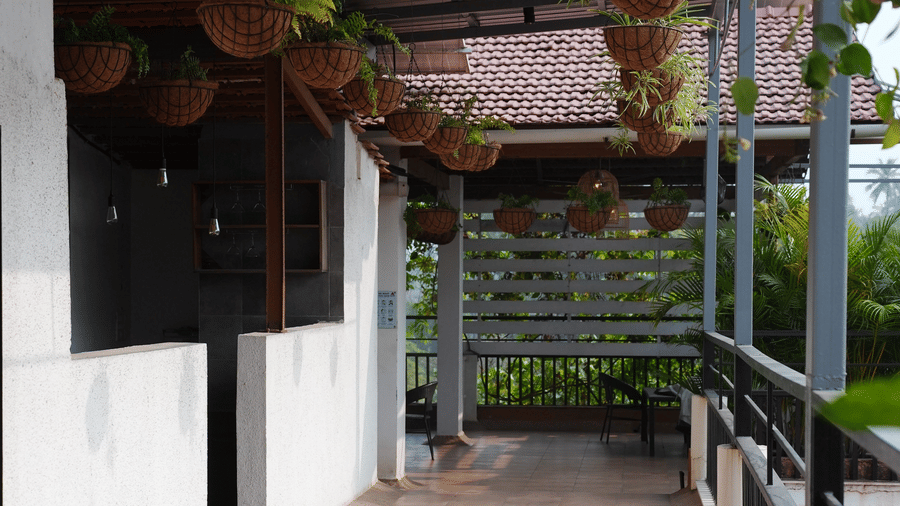 Hanging planters, potted plants, and a tiled walkway sit beneath a covered roof on a balcony at Bougainvillea, Alibaug.