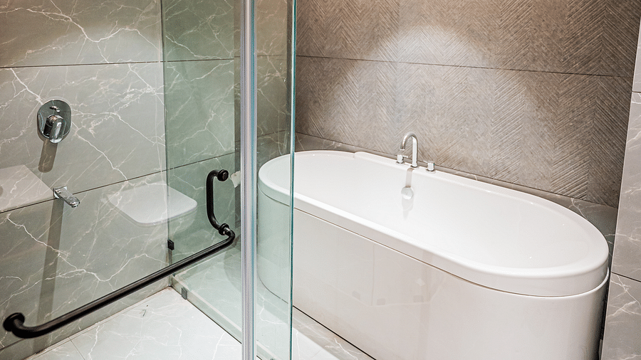 A spacious washroom at Indie Stays Mumbai International Airport features a bathtub with a glass panel separating the space, all with bright lighting.