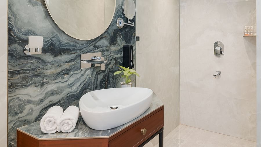 A spacious bathroom with wooden finishes that includes a wash basin and a wall-mounted round mirror | Hotel Ganga Maiya