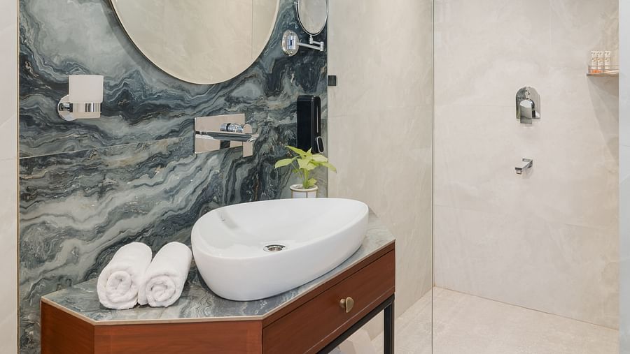 A spacious bathroom with wooden finishes that includes a wash basin and a wall-mounted round mirror | Hotel Ganga Maiya