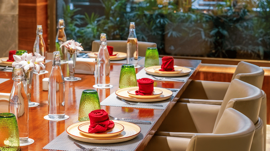 Napkins made into flowers on the plates set in a row at a table at Pushkara Resort & Spa by a glass window under warm lighting.