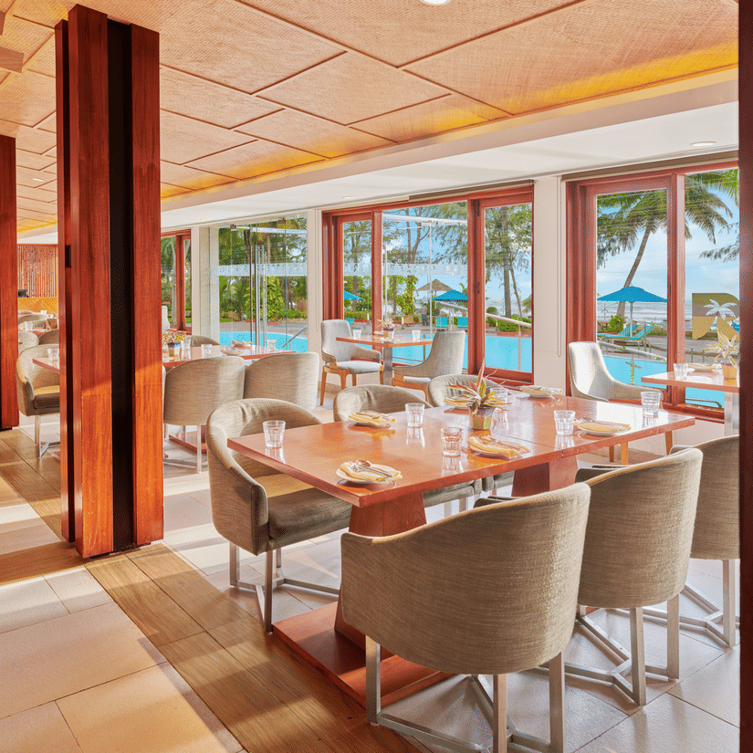 Chairs and tables placed right beside a glass window in a restaurant overlooking the swimming pool outside | The Resort, Mumbai