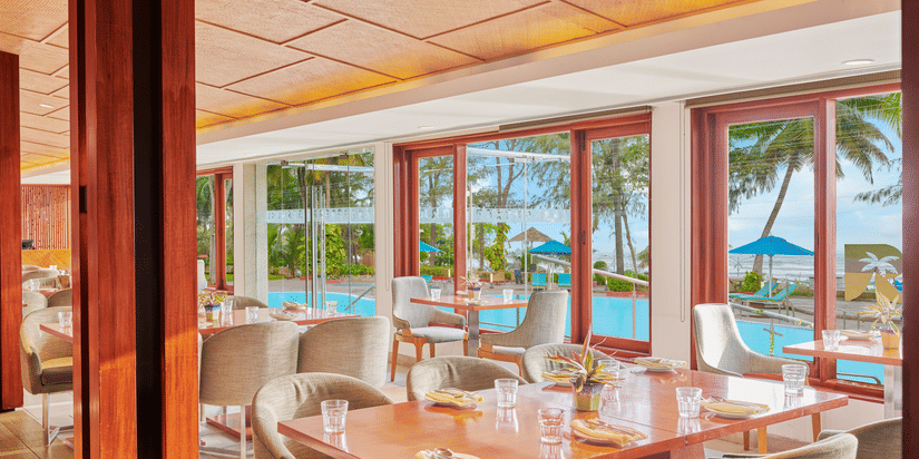 Chairs and tables placed right beside a glass window in a restaurant overlooking the swimming pool outside | The Resort, Mumbai