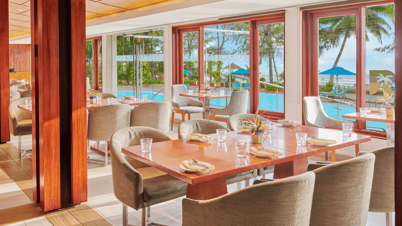 Chairs and tables placed right beside a glass window in a restaurant overlooking the swimming pool outside | The Resort, Mumbai