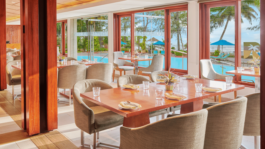 Chairs and tables placed right beside a glass window in a restaurant overlooking the swimming pool outside | The Resort, Mumbai