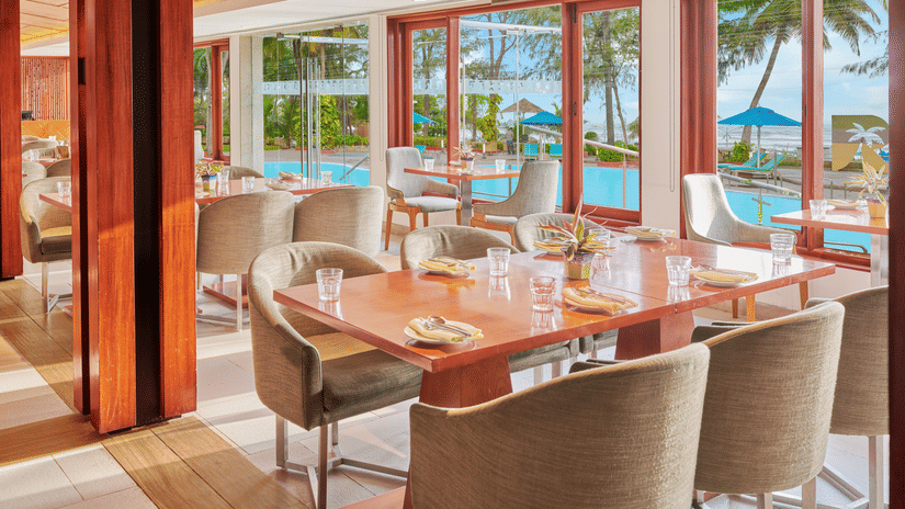 Chairs and tables placed right beside a glass window in a restaurant overlooking the swimming pool outside | The Resort, Mumbai