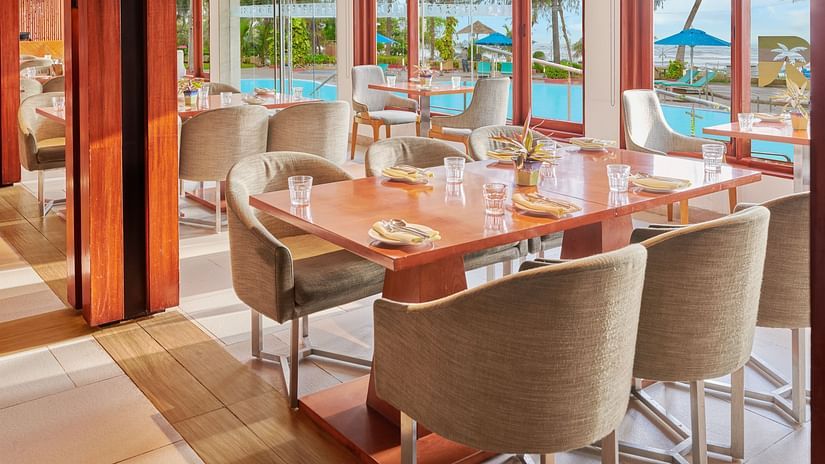 Chairs and tables placed right beside a glass window in a restaurant overlooking the swimming pool outside | The Resort, Mumbai