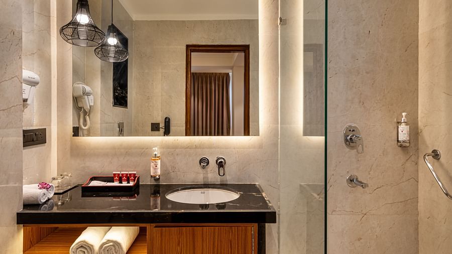 A modern bathroom with a glass-enclosed shower, dark countertop, and a large mirror with lighting. It features a wooden cabinet with open shelves, hanging pendant lights, and a partially visible white toilet - Lords Eco Inn Dehradun.