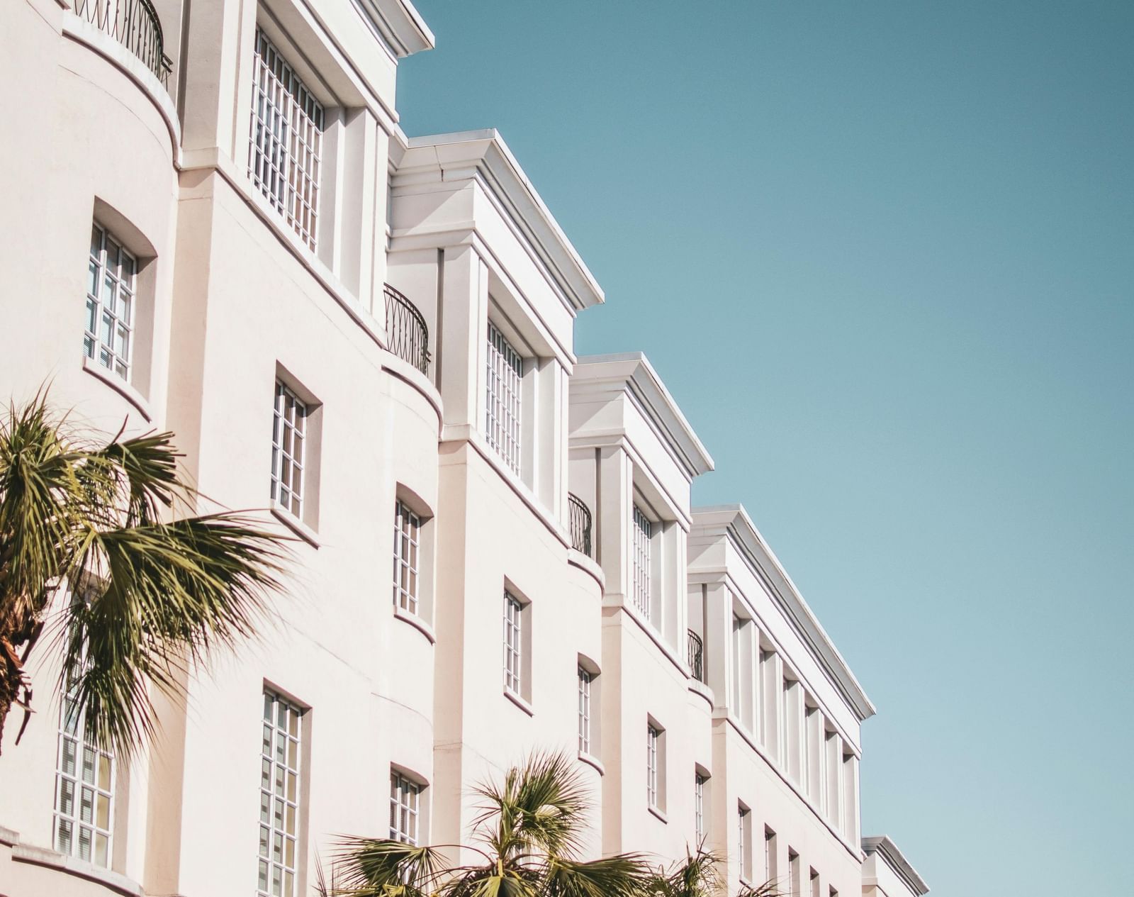 A low-angle shot of a white building with balconies and palm trees against a clear blue sky.