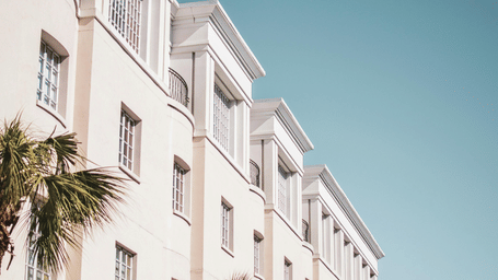 A low-angle shot of a white building with balconies and palm trees against a clear blue sky.