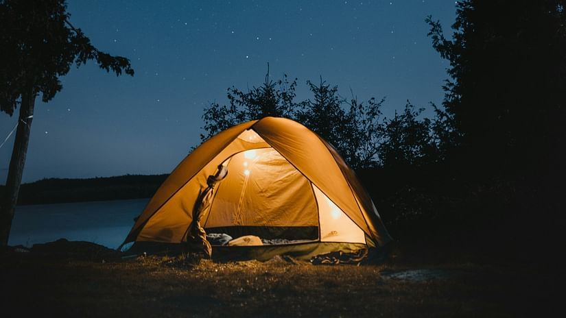 A glowing tent illuminated from within at night, nestled between silhouetted trees against a dark blue sky.