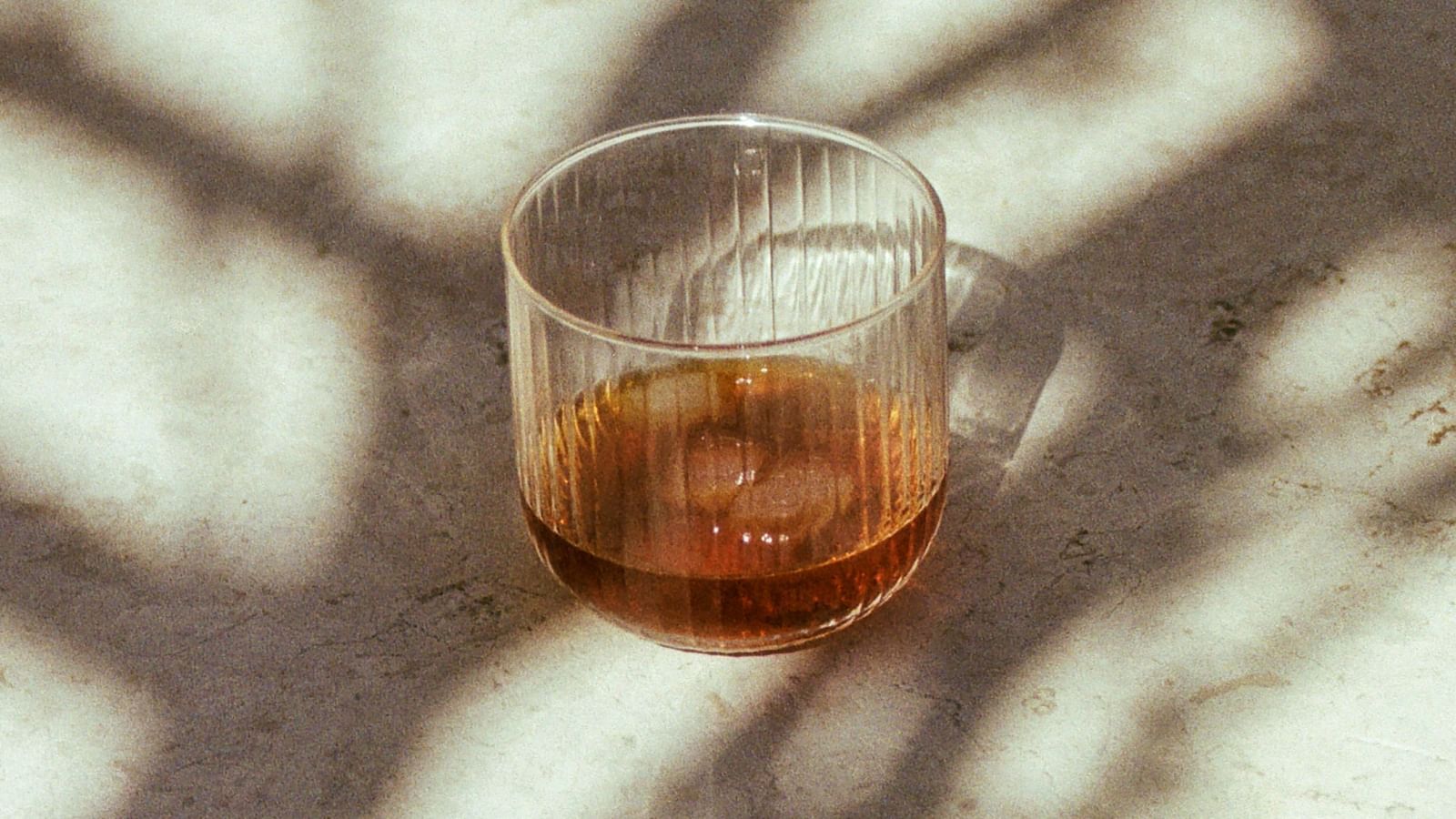 A solitary glass kept on a table filled with whisky and a tree branch's shadow falling on the glass.