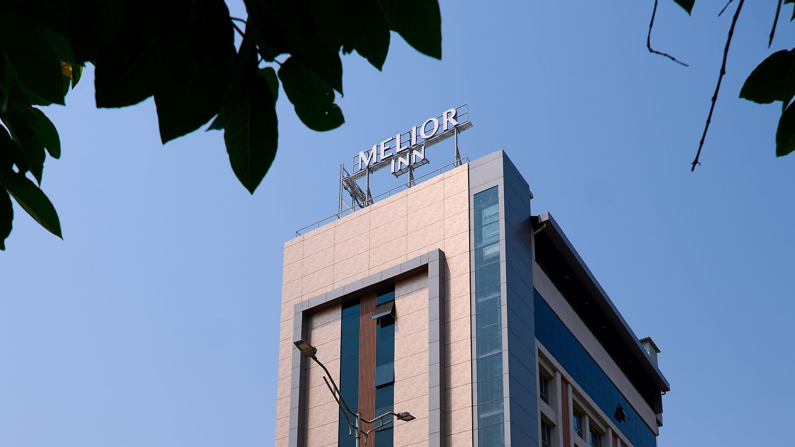 Angled street view of the hotel building, showing the full facade of the hotel framed by green tree branches and a sunny, cloudless sky.
