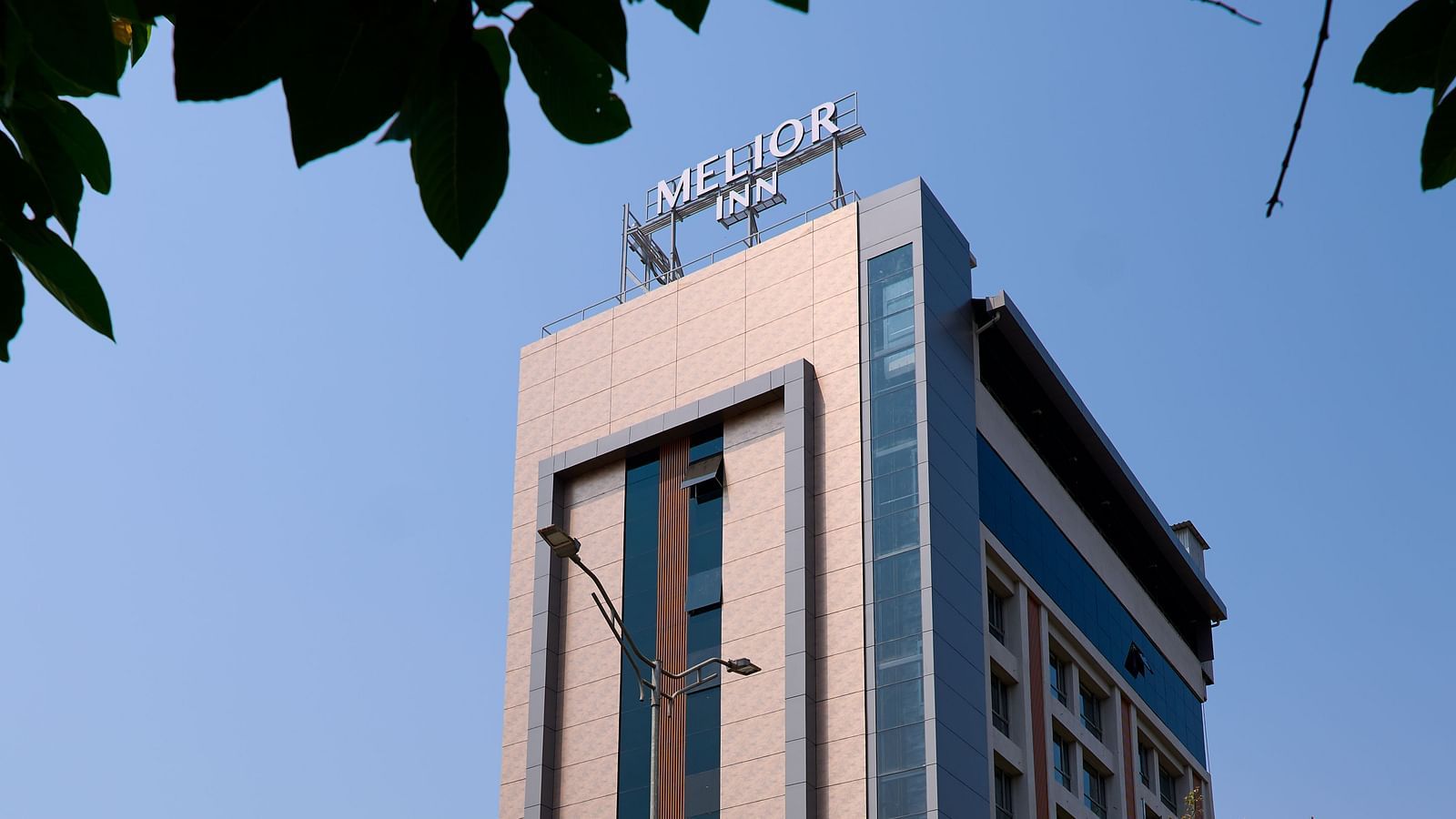 Angled street view of the hotel building, showing the full facade of the hotel framed by green tree branches and a sunny, cloudless sky.