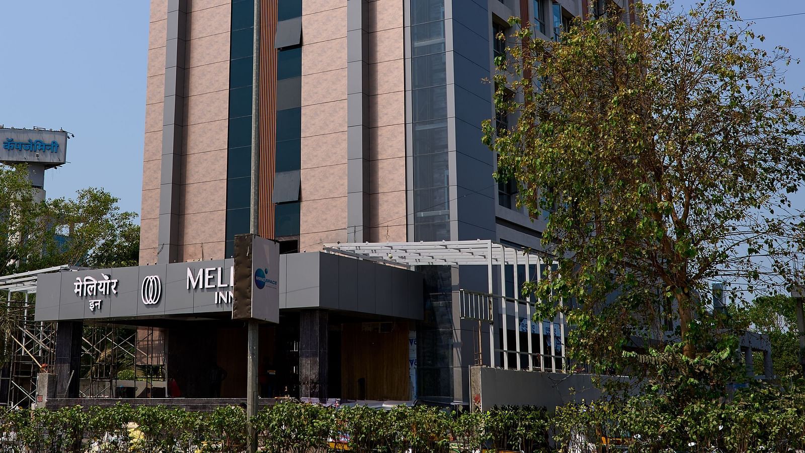 Angled street view of the hotel building, showing the full facade of the hotel framed by green tree branches and a sunny, cloudless sky.