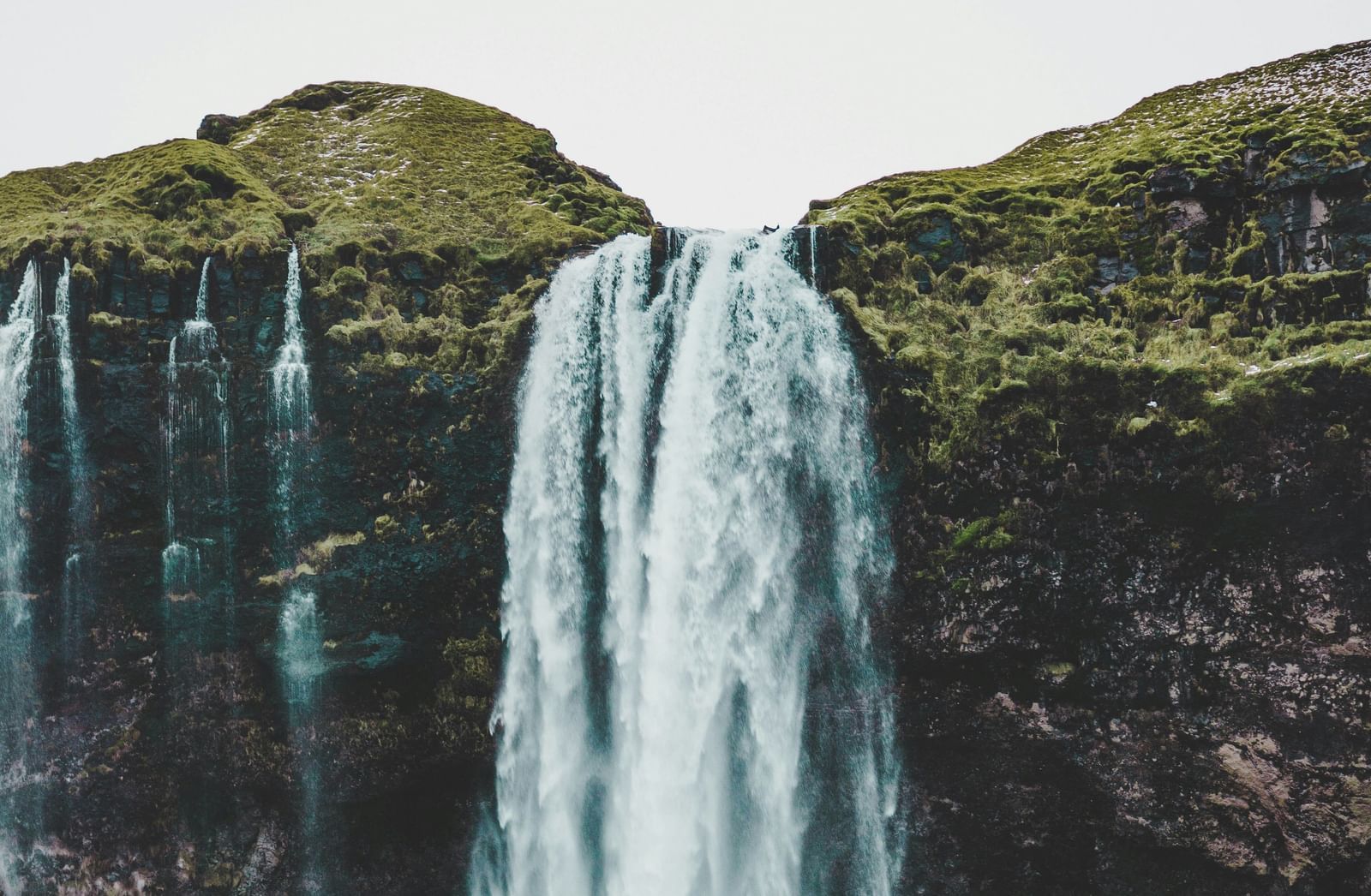 A majestic waterfall cascading down a lush, green cliff face, with tiny figures of people visible at the base, dwarfed by the immense natural beauty.