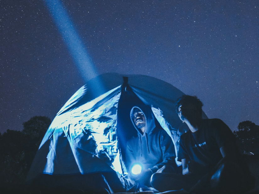 Illuminated camping tent beneath a clear night sky, with a bright torch beam pointing upwards across a peaceful mountain landscape
