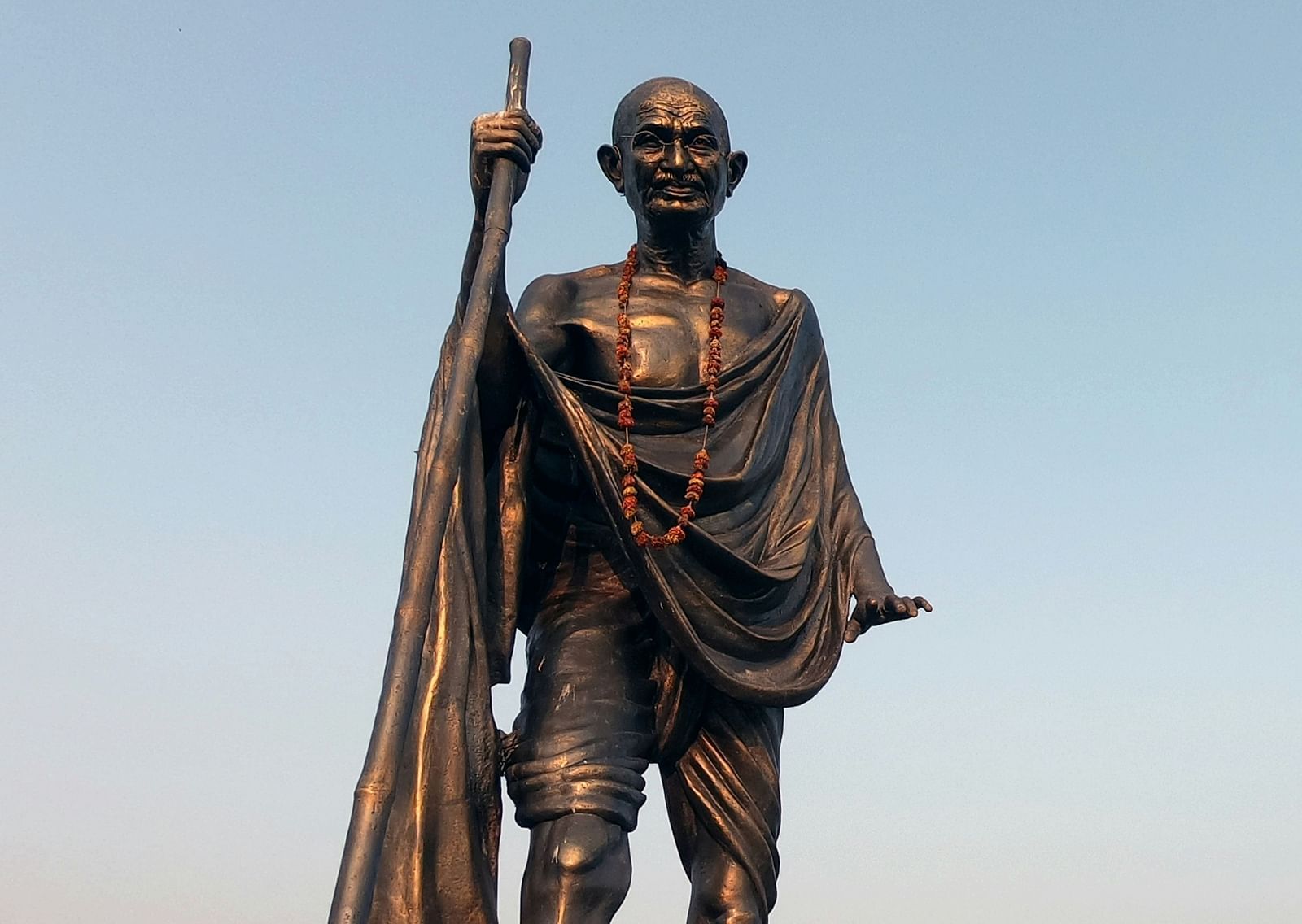 An overview of Mahathma Gandhi's statue holding a stick with blue sky in the background.