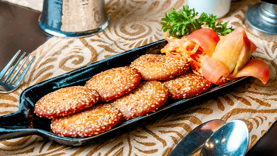A serving of golden-brown kebabs on a black platter at Za-aiqa, Noor-Us-Sabah Palace, Bhopal, garnished with fresh greens and tomato, presented on a table with an ivory patterned runner.