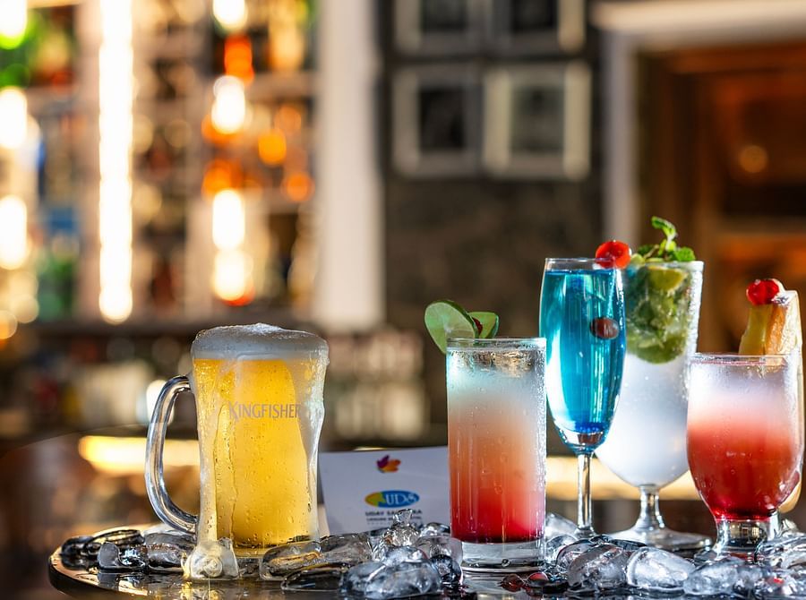 A selection of colourful cocktails and a frothy beer mug, garnished with cherries, lime, and mint, is placed on a reflective surface with ice cubes, set against a blurred pub background featuring the sign REUNION at Uday Samudra Leisure Beach Hotel.