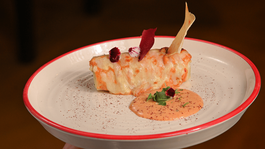 A person holding a white plate with a red rim, featuring an elegantly plated dish that includes a gourmet seafood item, garnishes, and a dollop of creamy orange sauce - Ananta Spa and Resort, Pushkar.