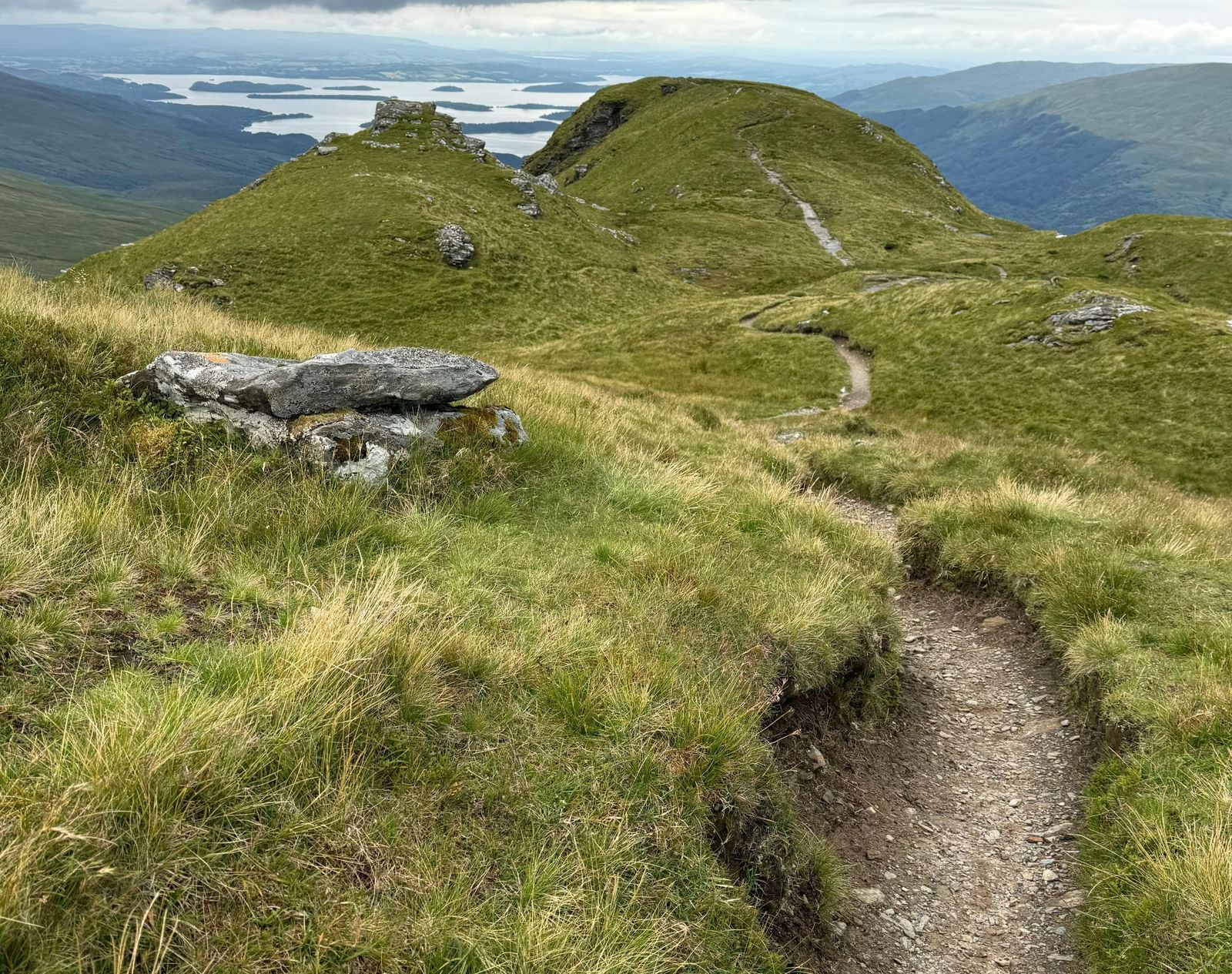 A rugged dirt trail meanders through lush green hills, leading towards distant peaks under a dramatic, overcast sky with patches of light on the horizon.