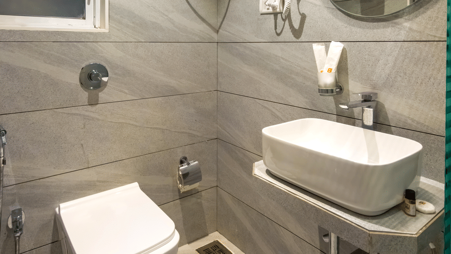 Modern bathroom with a western toilet, ceramic vessel sink, round mirror, and wall-mounted hairdryer in the Deluxe Room at Home2 Residency, Mumbai Airport.