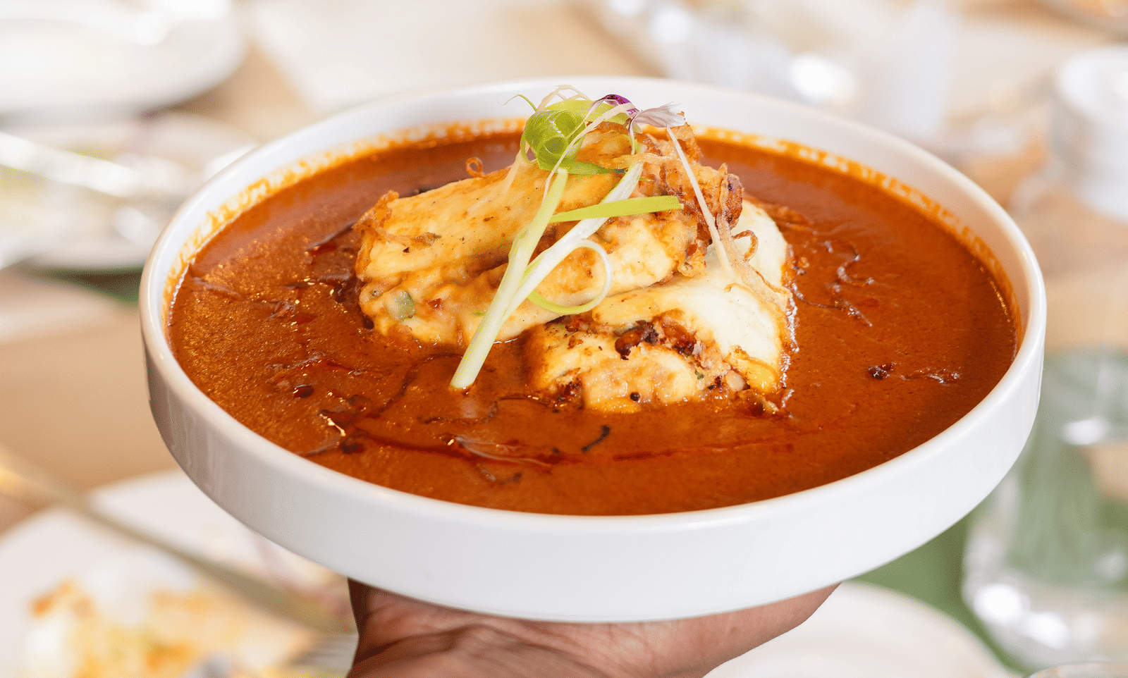 A person is presenting a large, white bowl of rich, copper-coloured curry at a busy table - Megha Resort & Villas, Dehradun