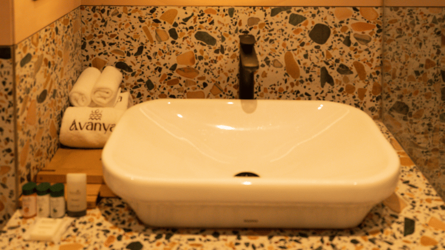 A white ceramic washbasin sits on a multi-coloured terrazzo countertop beneath a circular illuminated mirror at Avanya Resorts N Retreat.