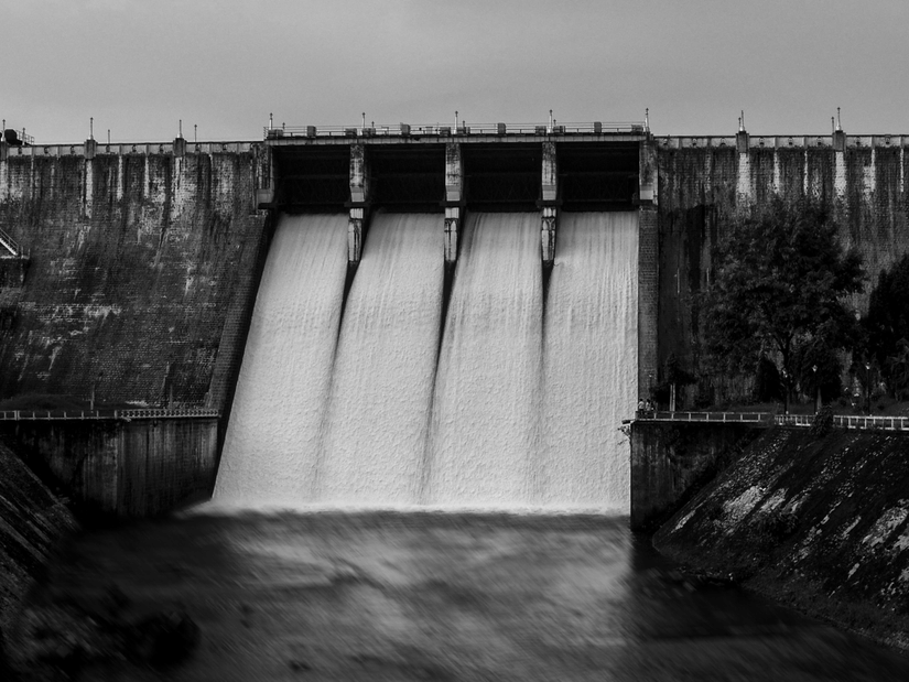 A black and white photograph of a large concrete dam with water rushing through its spillway gates.