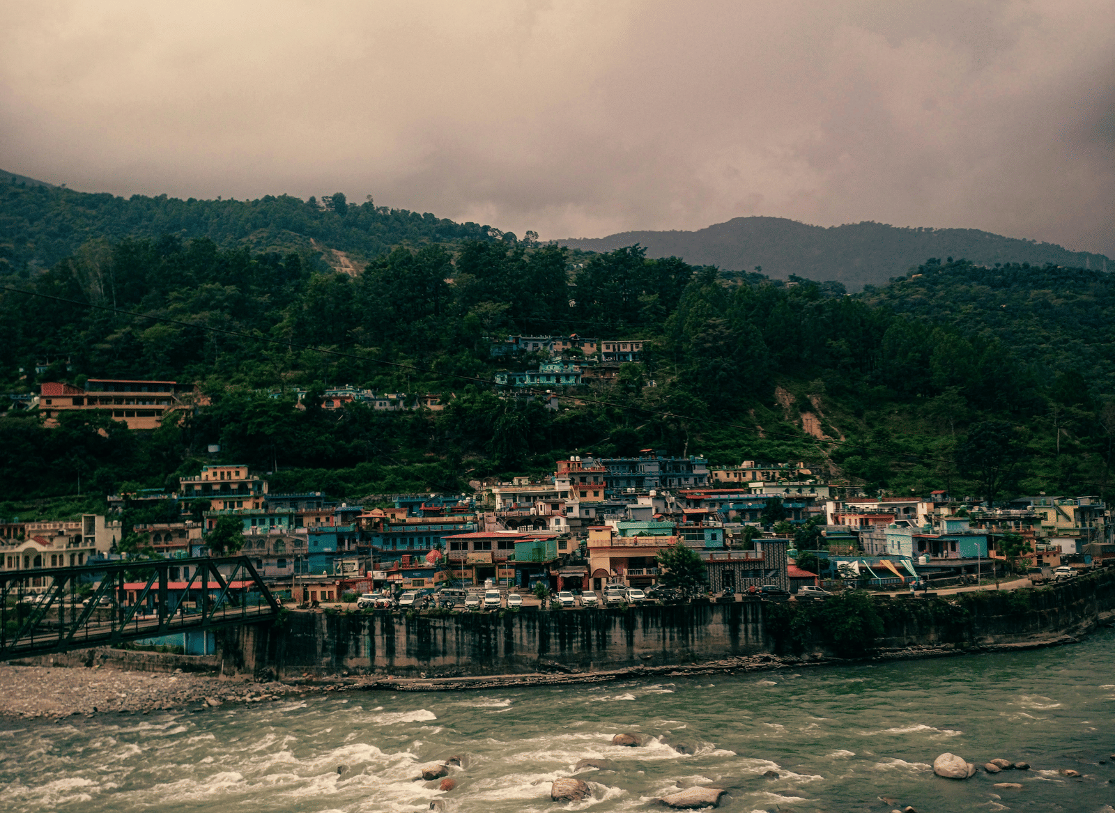 Cloudy day view of a small town with buildings nestled beside a river and forested mountains.