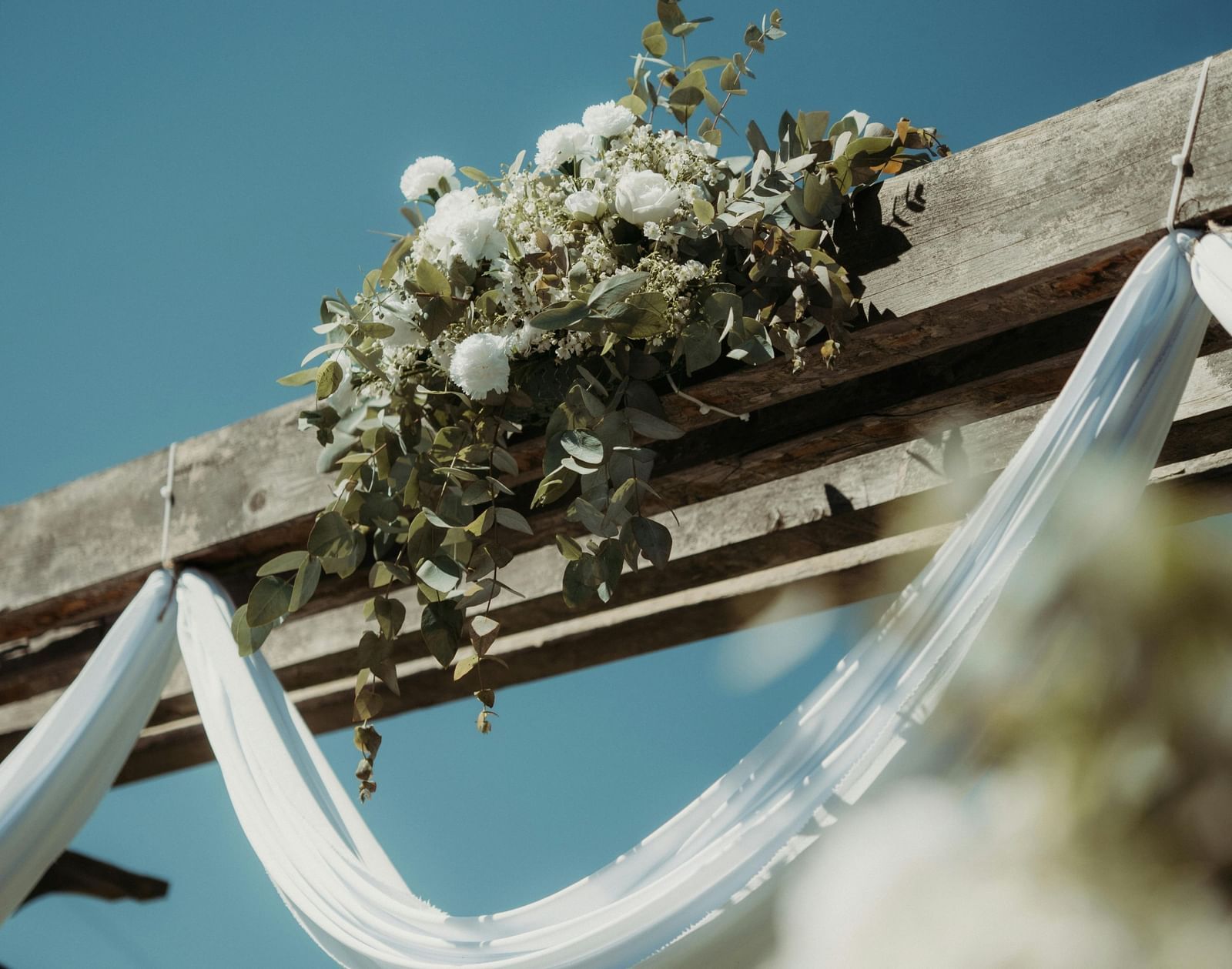  A wooden arch decorated with white flowers and draped with white fabric, set against a clear blue sky at The Groveland Hotel.
