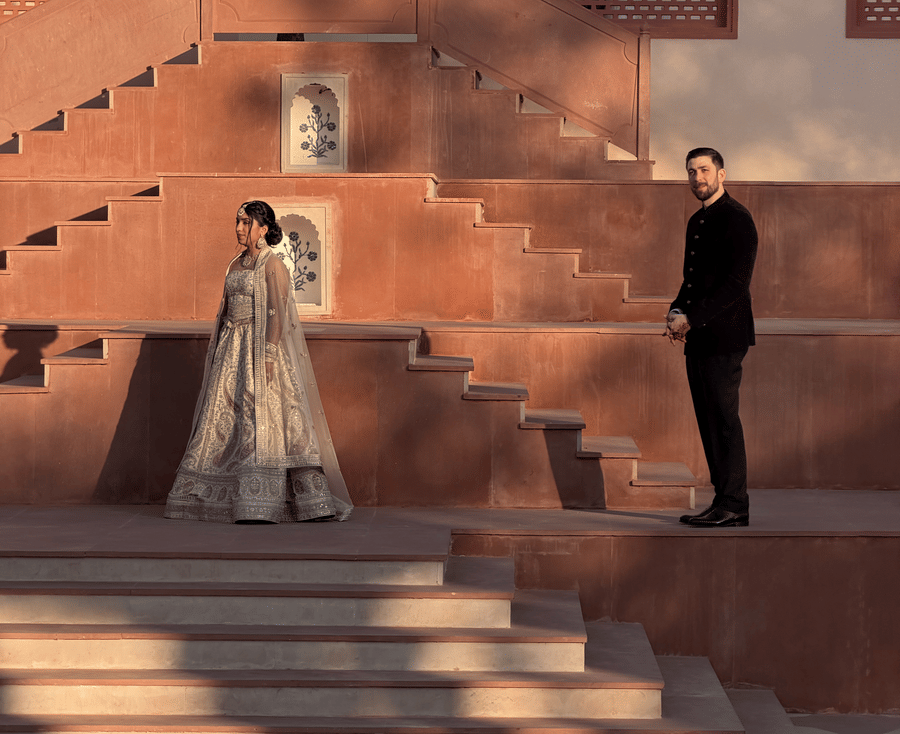A bride stands on the wide stone steps while the groom looks up at her from below against the traditional red-stone architecture of Umaid Palace.
