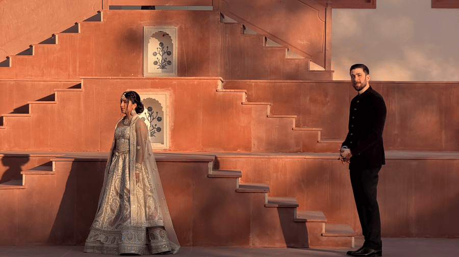 A bride stands on the wide stone steps while the groom looks up at her from below against the traditional red-stone architecture of Umaid Palace.