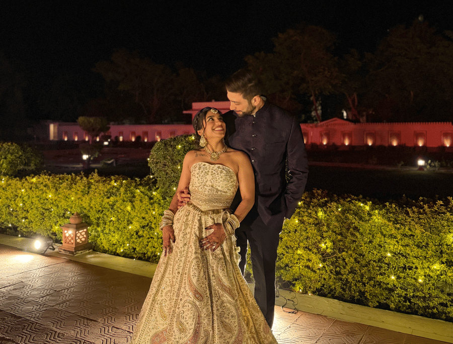 A bride and groom posing together on an outdoor lawn at night with the illuminated structure of Umaid Palace visible in the background.